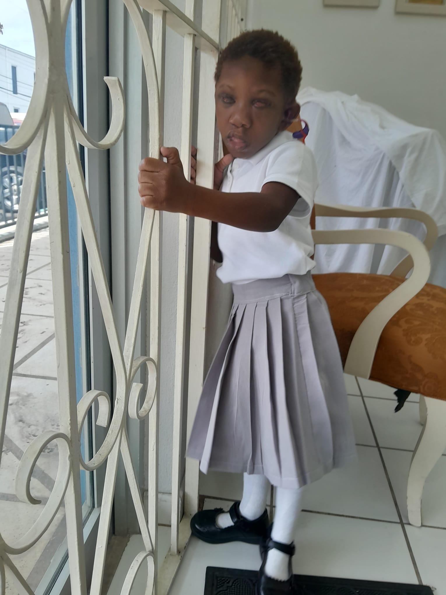 Ja’Dei Grant standing in her white-and-grey school uniform and holding a decorative white security grille at a glass doorway, with a cushioned chair behind her.