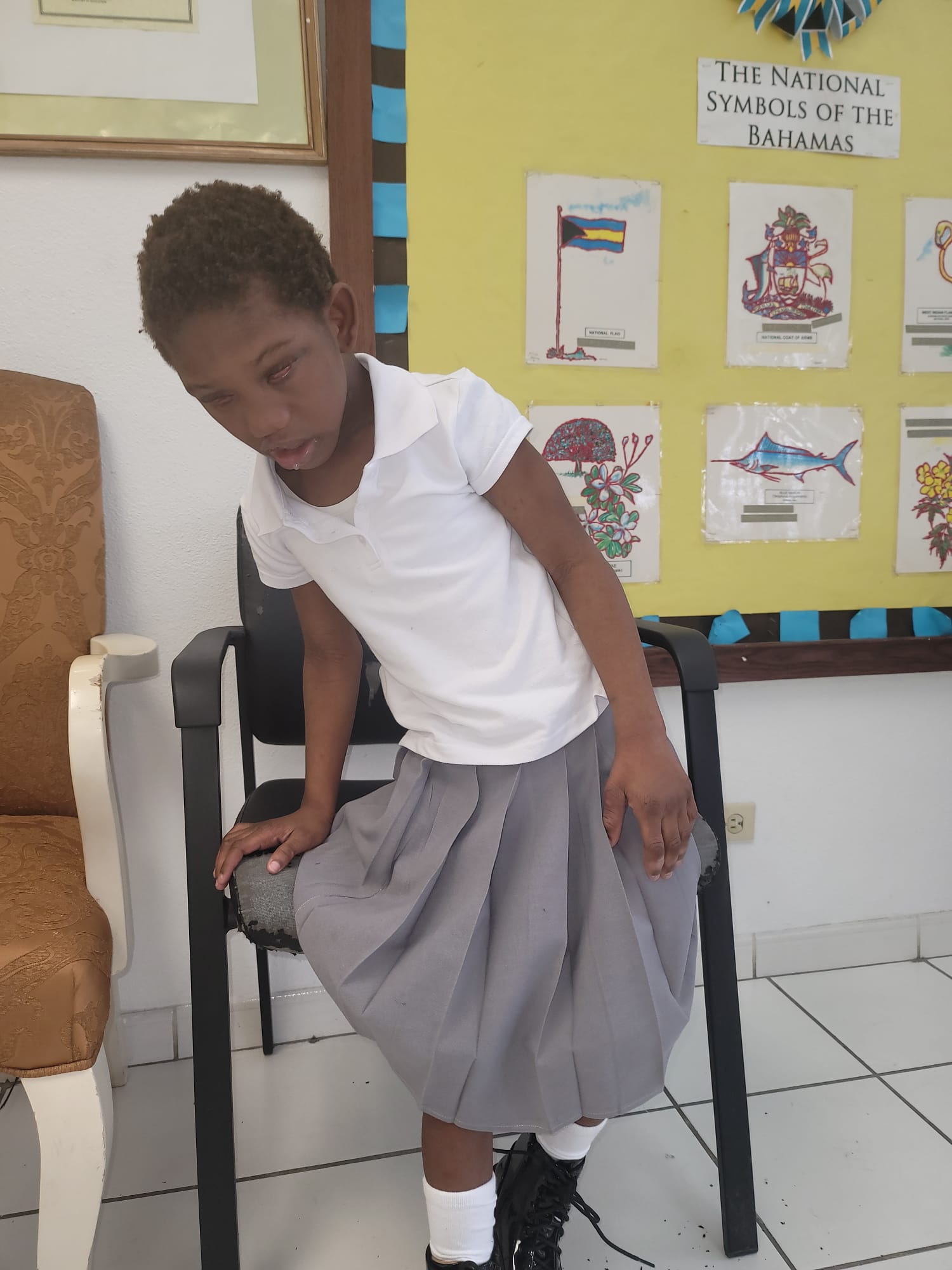 Ja’Dei Grant standing beside a black chair at school in front of a bulletin board displaying the national symbols of The Bahamas.