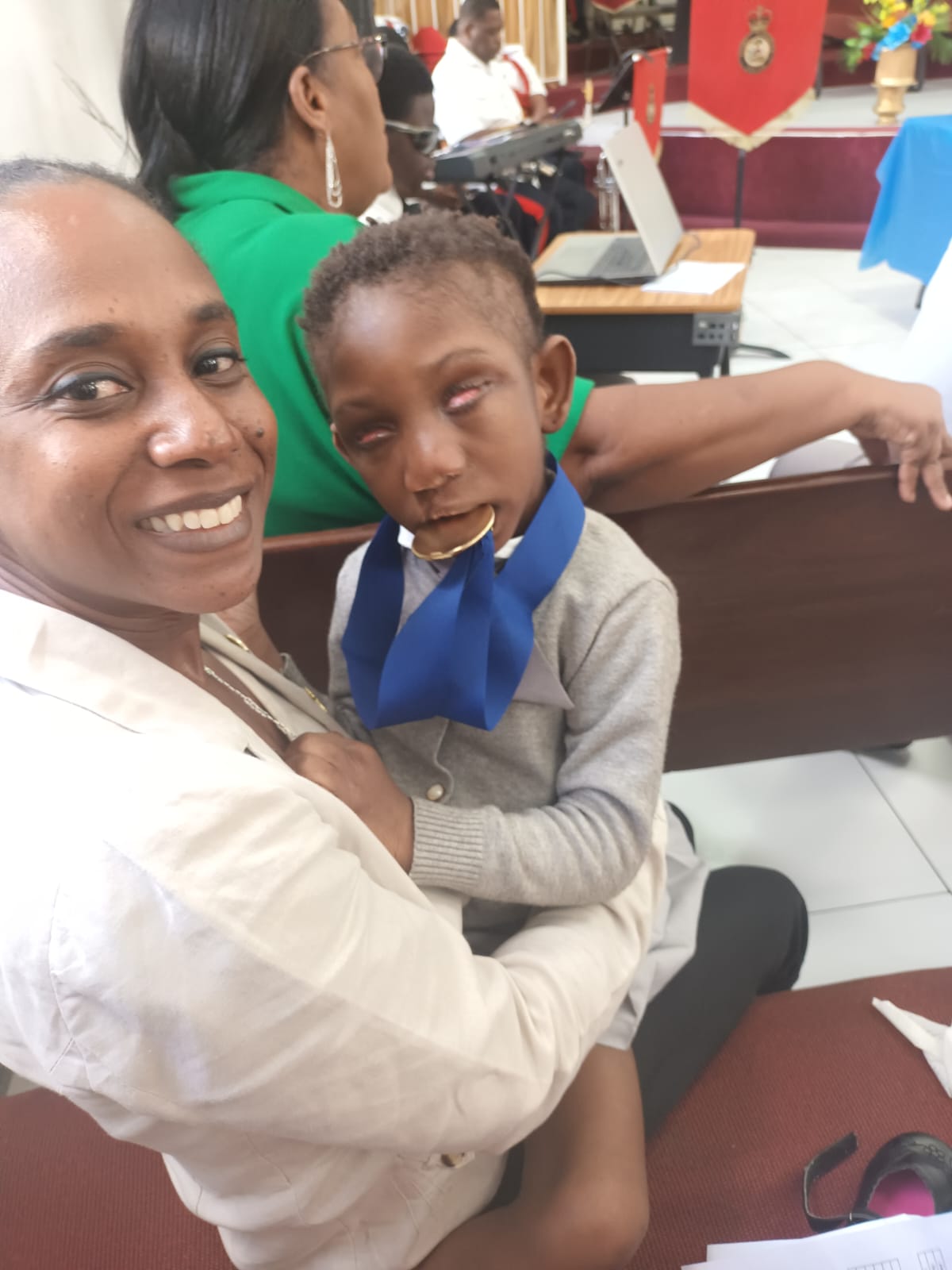 A close photograph of Ja’Dei Grant being held on a smiling woman’s lap during a celebration, wearing a grey cardigan and a large blue ribbon around her neck.