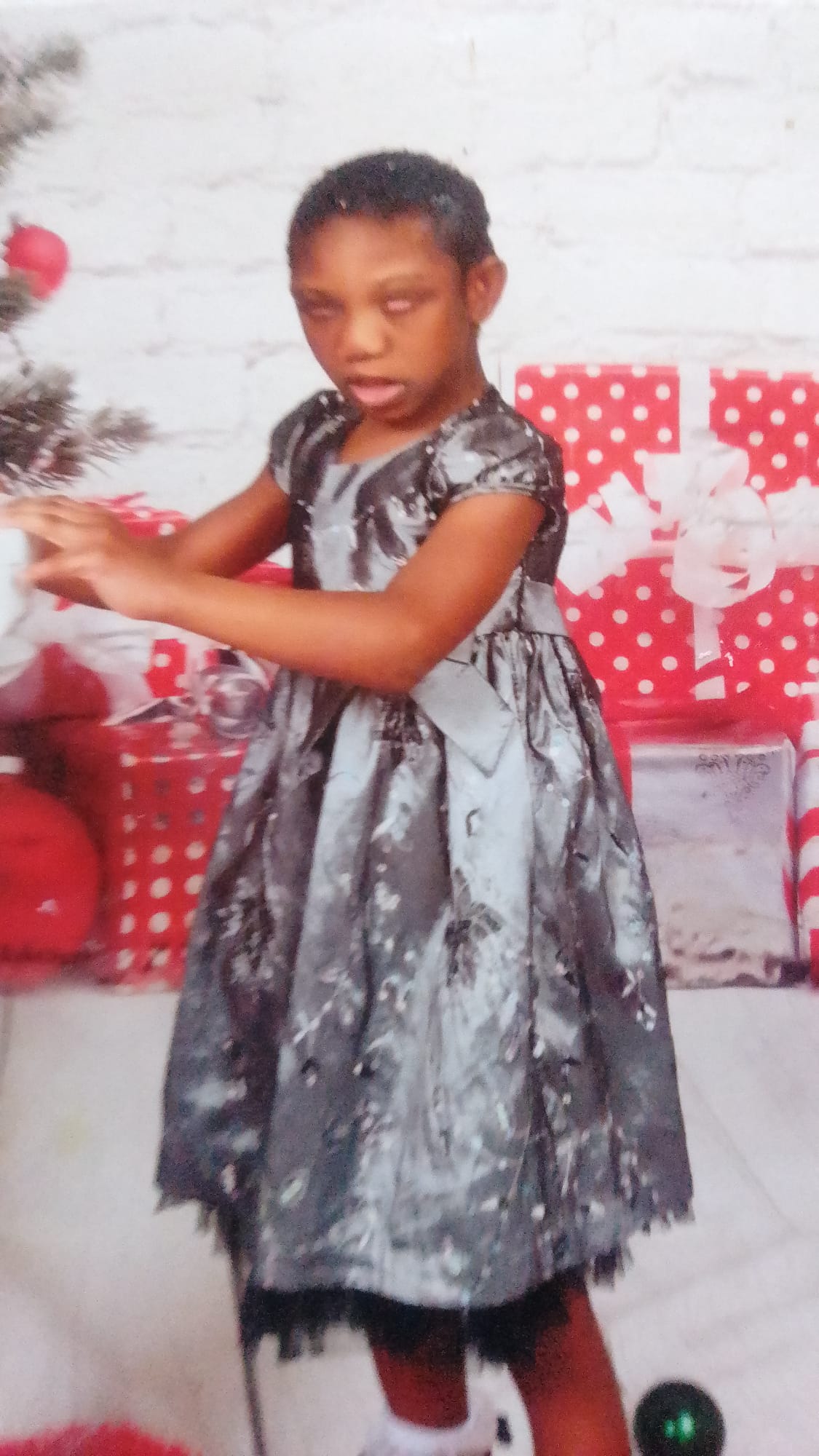 A closer photograph of Ja’Dei Grant in her black, silver and grey party dress, with her arms extended beside a Christmas tree and red-and-white presents.