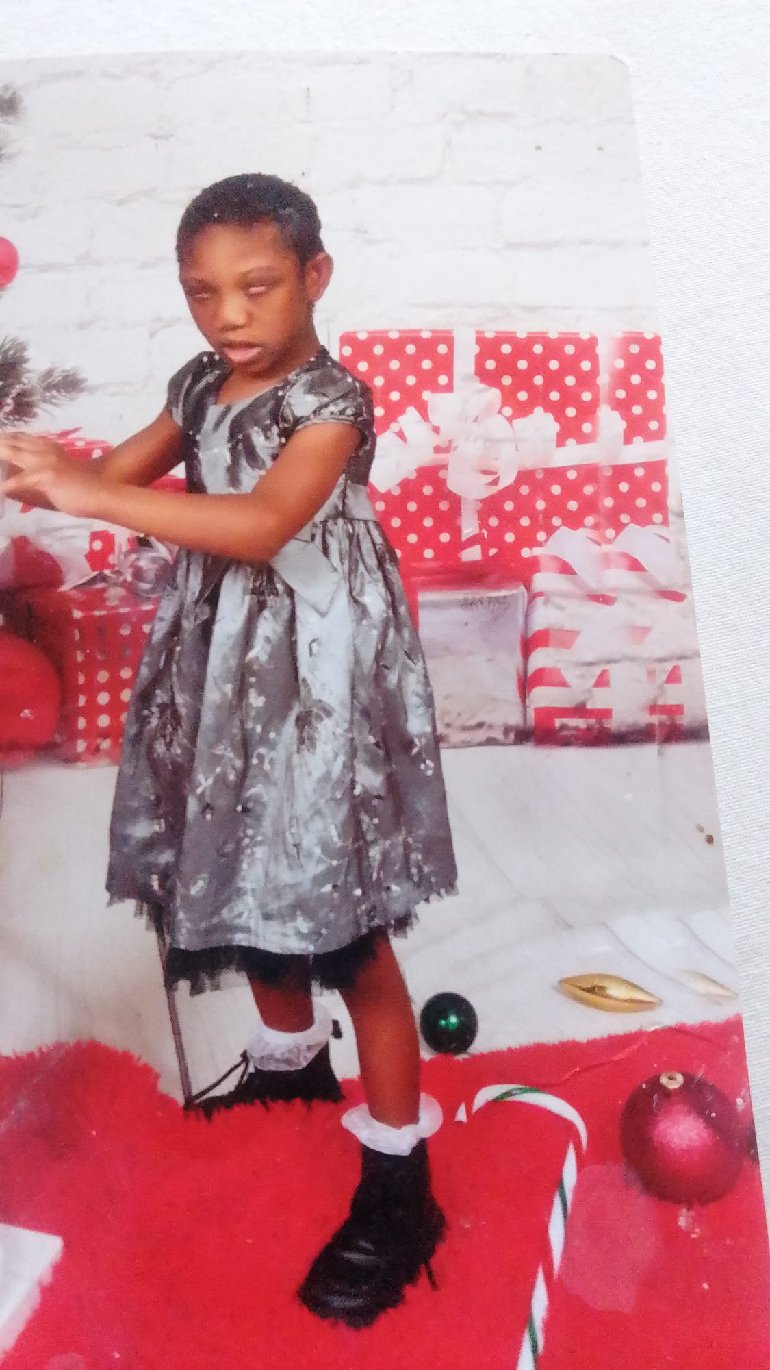 Ja’Dei Grant standing beside a Christmas tree in a black, silver and grey party dress, surrounded by large red-and-white wrapped presents and festive decorations.