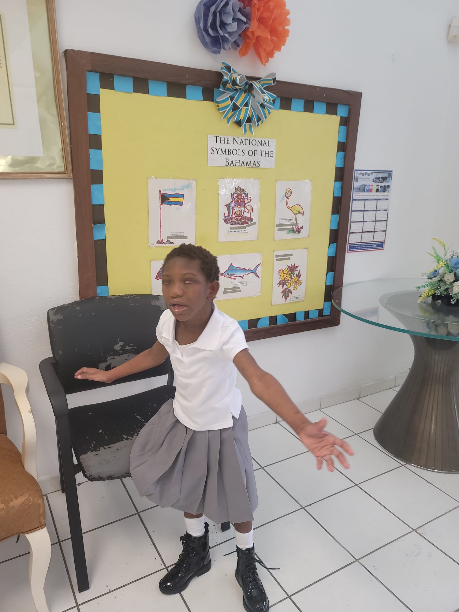Ja’Dei Grant standing at school in her white-and-grey uniform with her arms extended outward. Behind her is a colourful bulletin board displaying the national symbols of The Bahamas.