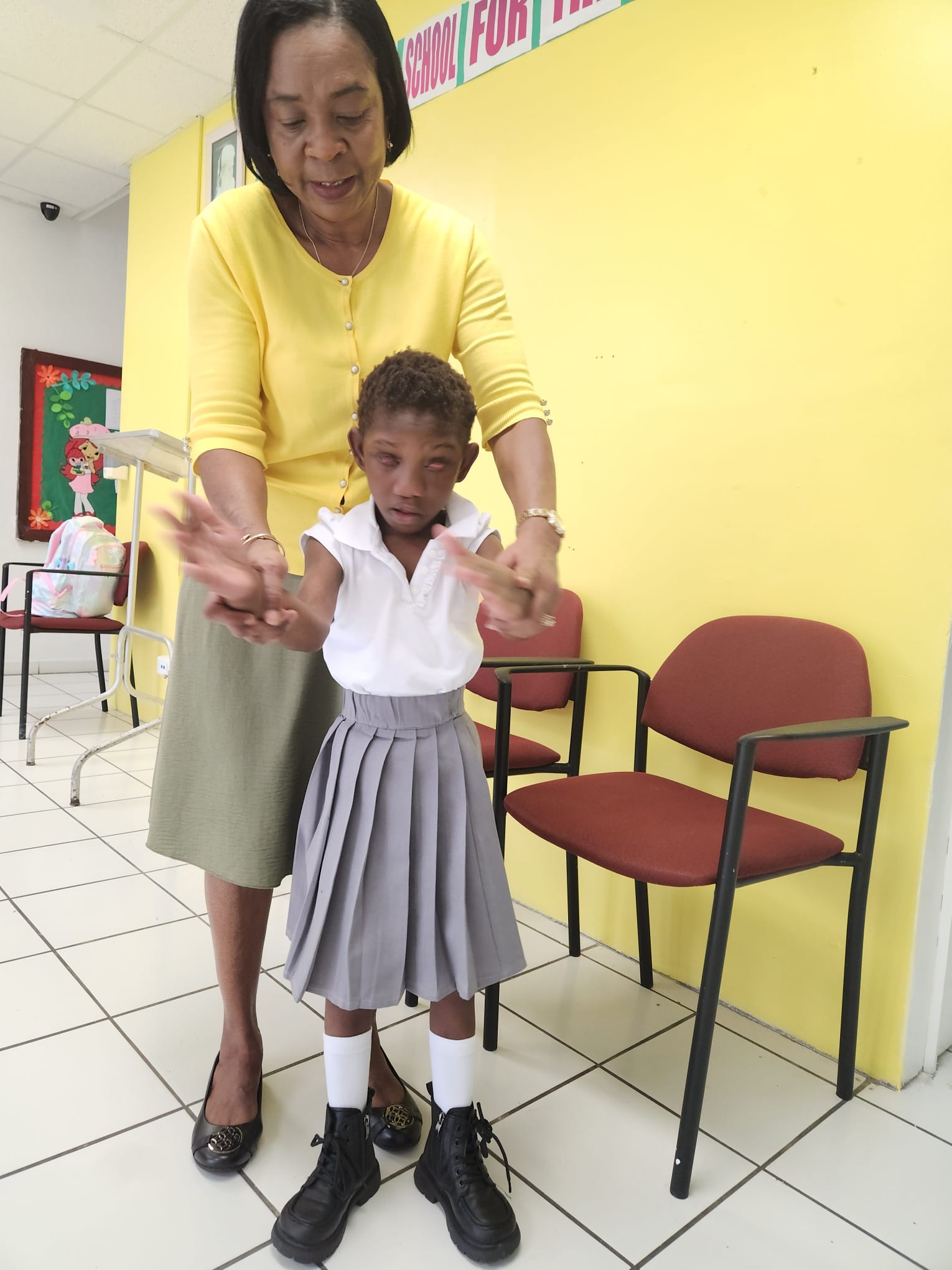 Ja’Dei Grant standing in her school uniform while a woman in a yellow cardigan gently supports her forearms and guides both hands forward during a movement activity.
