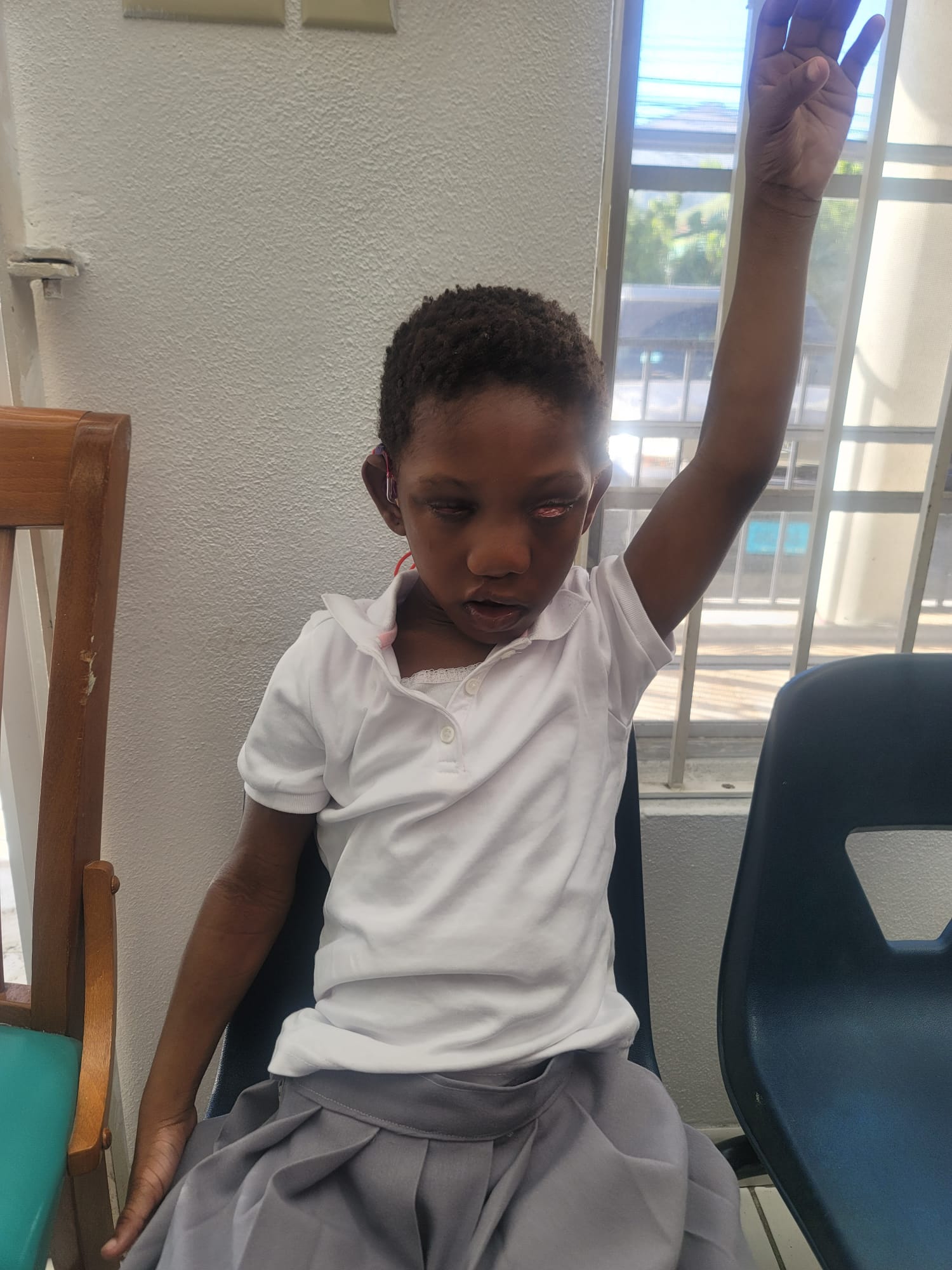 Ja’Dei Grant seated near a bright window in her white-and-grey school uniform, raising one arm high above her head with her palm open.