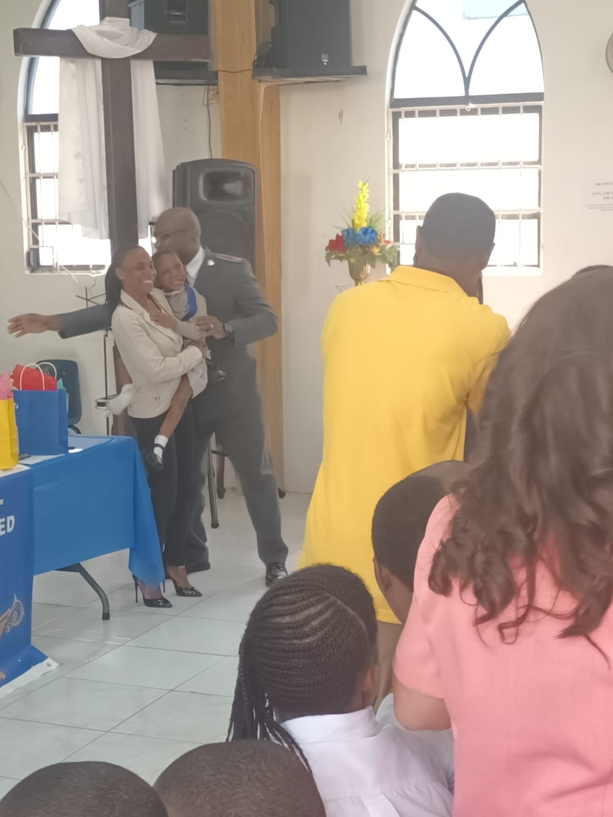 Ja’Dei Grant being lovingly held by a smiling woman during a school or church celebration, with a uniformed Salvation Army officer standing closely behind them.